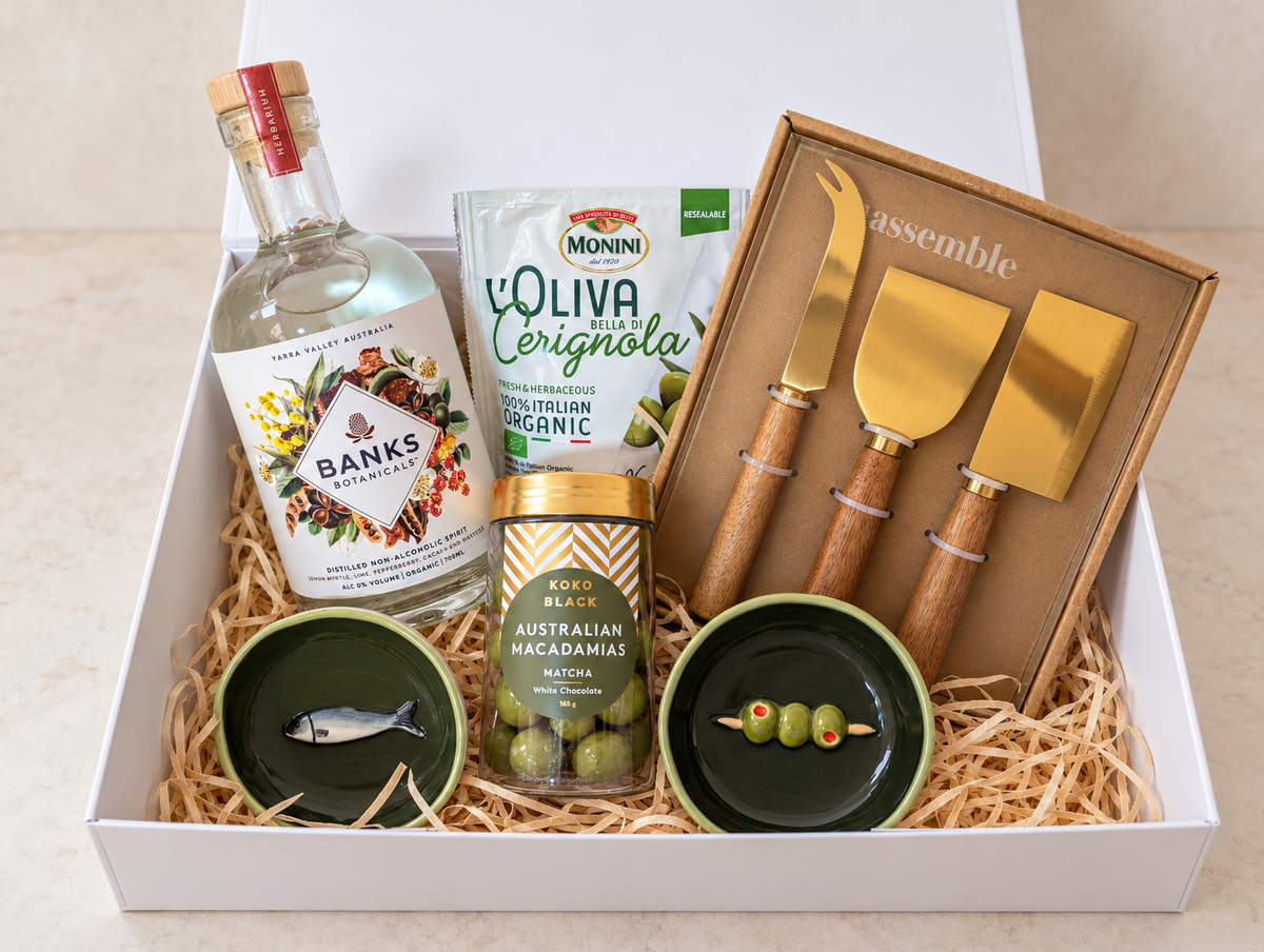 Non-alcoholic Botanical Gathering Gift Box with Banks Botanicals, olives, matcha macadamias, three cheese knives and two green ceramic dishes.